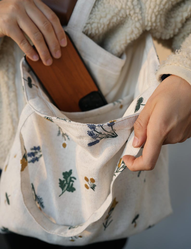 May include: Cream-colored tote bag with a floral pattern. The bag has a pocket and a wooden phone case is visible inside. The floral design includes green, yellow, and blue botanical illustrations. The bag is held by a person.
