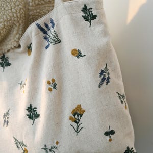 May include: Cream-colored tote bag with embroidered floral patterns in yellow, green, and blue. The bag features a simple design with a rectangular shape and long straps. The floral embroidery adds a touch of nature-inspired charm to the bag.