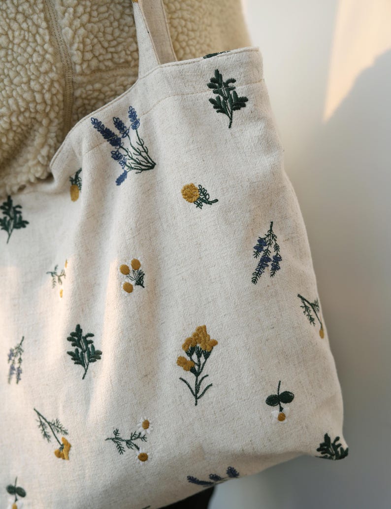 May include: Cream-colored tote bag with embroidered floral patterns in yellow, green, and blue. The bag features a simple design with a rectangular shape and long straps. The floral embroidery adds a touch of nature-inspired charm to the bag.