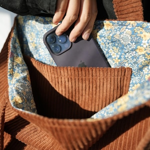 May include: A brown corduroy tote bag with a floral patterned lining. A dark gray smartphone is in the front pocket. The bag is made of a ribbed fabric and has a contrasting floral interior.