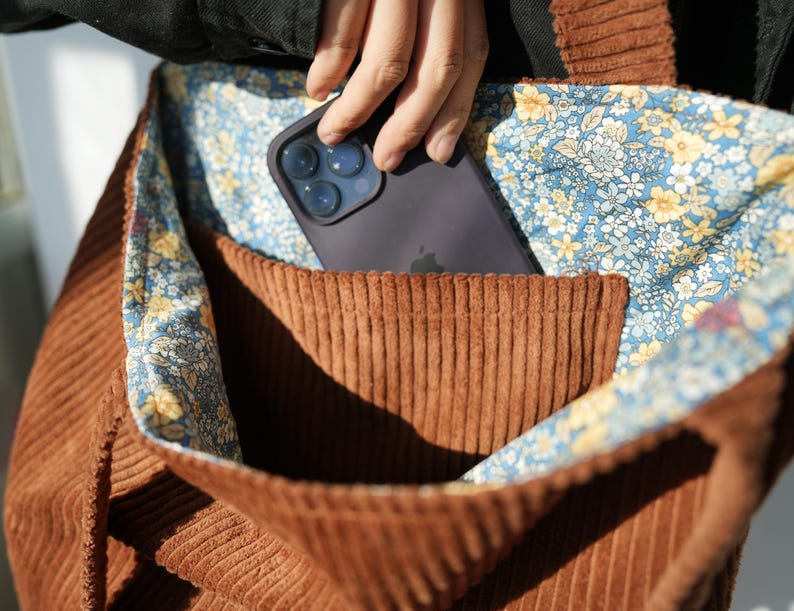 May include: A brown corduroy tote bag with a floral patterned lining. A dark gray smartphone is in the front pocket. The bag is made of a ribbed fabric and has a contrasting floral interior.