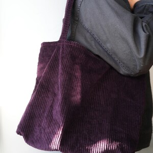 Op de afbeelding: Een dieppaarse corduroy tote bag met een brede schouderriem. De tas is gemaakt van een geribbelde stof en heeft een eenvoudige, rechthoekige vorm. De tas wordt over de schouder gedragen.