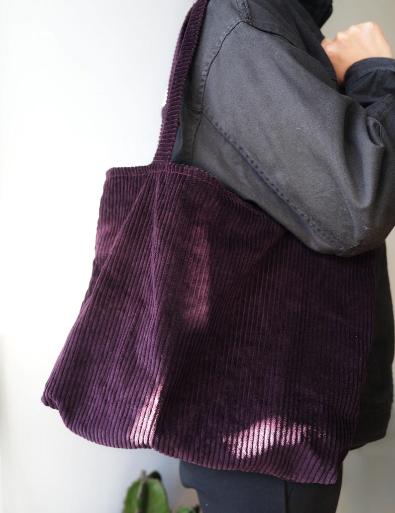 Op de afbeelding: Een dieppaarse corduroy tote bag met een brede schouderriem. De tas is gemaakt van een geribbelde stof en heeft een eenvoudige, rechthoekige vorm. De tas wordt over de schouder gedragen.