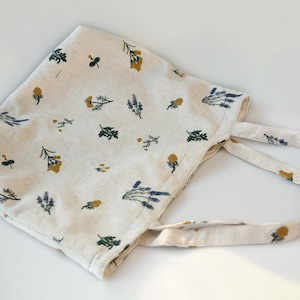 May include: Off-white tote bag with a floral pattern featuring yellow, green, and blue botanical illustrations. The bag has two handles and is made of a soft, textured fabric. The bag is designed for carrying items.