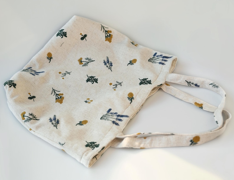 May include: Off-white tote bag with a floral pattern featuring yellow, green, and blue botanical illustrations. The bag has two handles and is made of a soft, textured fabric. The bag is designed for carrying items.