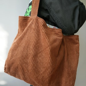 May include: A brown corduroy tote bag with vertical ribbing. The bag has two long straps and a rectangular shape. The bag is made of a soft, textured fabric and appears to be empty. The bag is being held by a person.