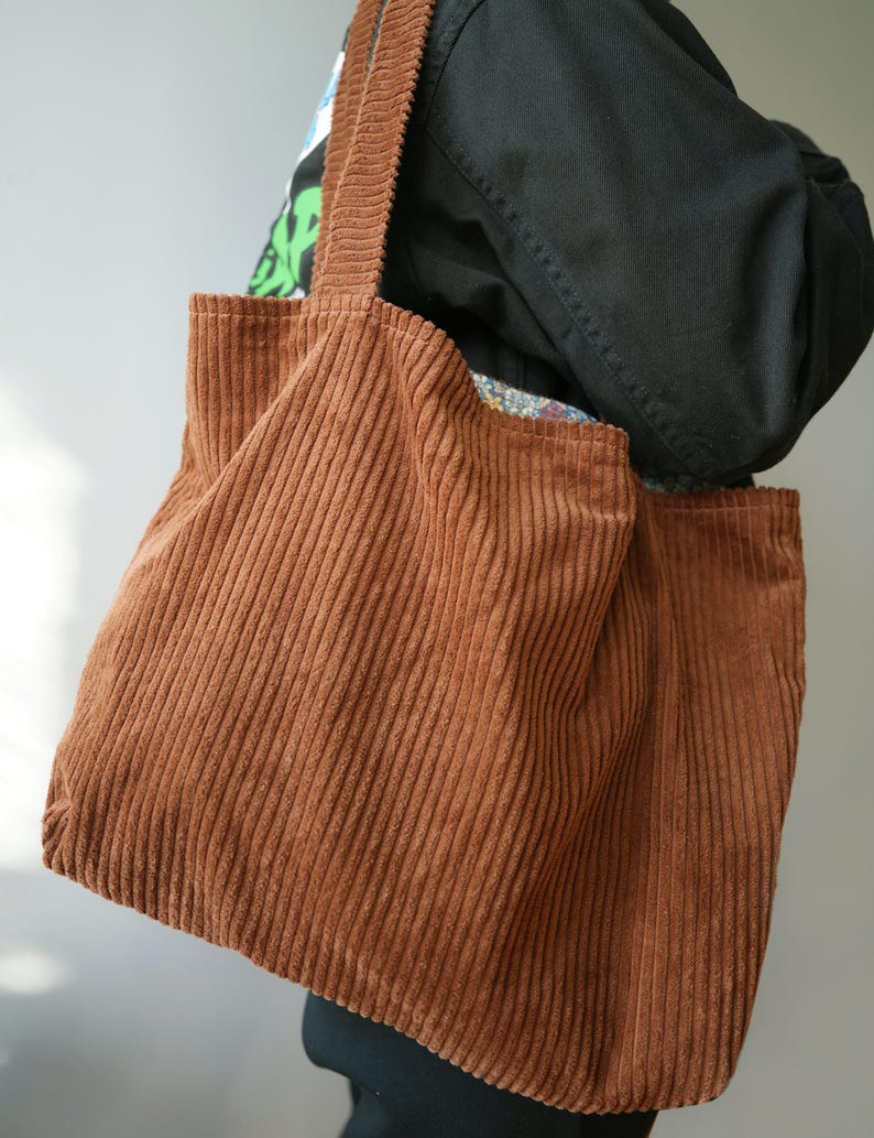 May include: A brown corduroy tote bag with vertical ribbing. The bag has two long straps and a rectangular shape. The bag is made of a soft, textured fabric and appears to be empty. The bag is being held by a person.
