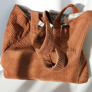 May include: A brown corduroy tote bag with matching handles. The bag has a rectangular shape and a floral patterned lining. The corduroy fabric has a ribbed texture. The bag is sitting on a white surface.