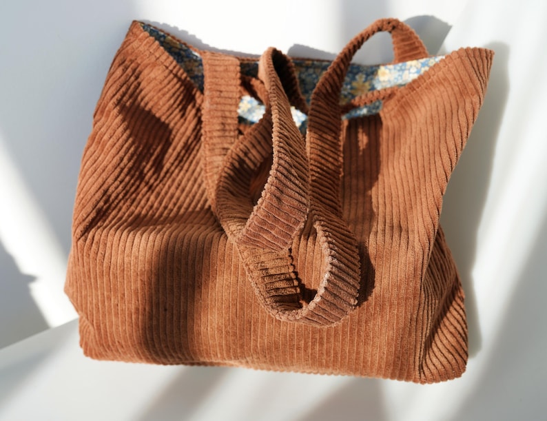 May include: A brown corduroy tote bag with matching handles. The bag has a rectangular shape and a floral patterned lining. The corduroy fabric has a ribbed texture. The bag is sitting on a white surface.
