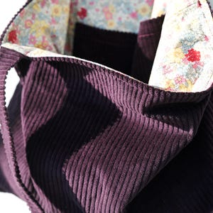 Op de afbeelding: Een dieppaarse corduroy tote bag met een bloemenvoering. De tas heeft een gestructureerde vorm met een brede opening en stevige handvatten. De binnenvoering heeft een kleurrijke bloemenprint met rode, roze en blauwe bloemen.