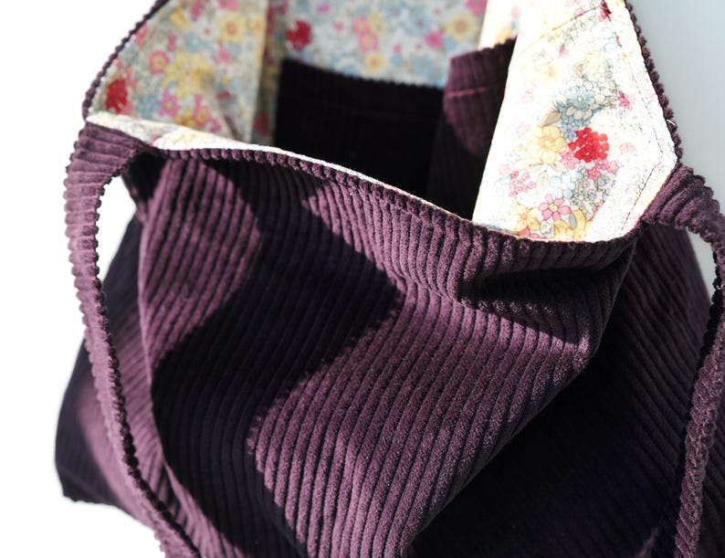 Op de afbeelding: Een dieppaarse corduroy tote bag met een bloemenvoering. De tas heeft een gestructureerde vorm met een brede opening en stevige handvatten. De binnenvoering heeft een kleurrijke bloemenprint met rode, roze en blauwe bloemen.