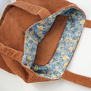 May include: A brown corduroy tote bag with a floral patterned lining. The bag has a rectangular shape with a wide opening and two long straps. The interior lining features a blue background with yellow and white flowers.