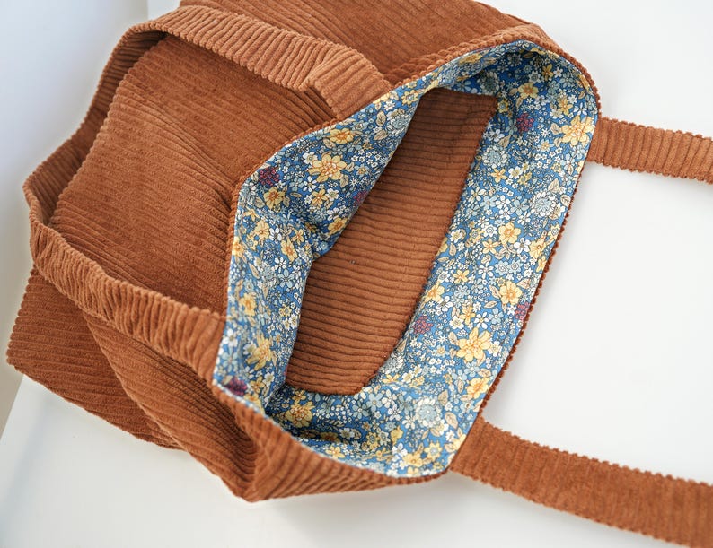 May include: A brown corduroy tote bag with a floral patterned lining. The bag has a rectangular shape with a wide opening and two long straps. The interior lining features a blue background with yellow and white flowers.