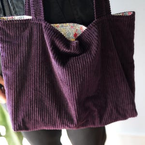 Op de afbeelding: Een dieppaarse corduroy tote bag met een bloemenpatroon voering. De tas heeft twee stevige handvatten en een brede opening. De corduroy stof heeft verticale ribbels, en de tas lijkt goed gemaakt.