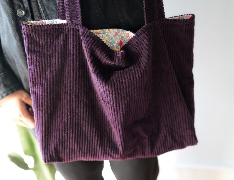 Op de afbeelding: Een dieppaarse corduroy tote bag met een bloemenpatroon voering. De tas heeft twee stevige handvatten en een brede opening. De corduroy stof heeft verticale ribbels, en de tas lijkt goed gemaakt.