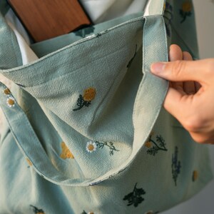May include: A light green tote bag with a floral pattern. The bag features embroidered yellow and white flowers, with a white interior. A wooden object is visible inside the bag's pocket. The bag is held open by a hand.