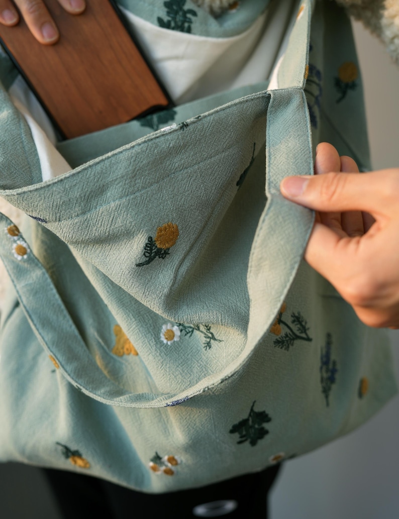 May include: A light green tote bag with a floral pattern. The bag features embroidered yellow and white flowers, with a white interior. A wooden object is visible inside the bag's pocket. The bag is held open by a hand.