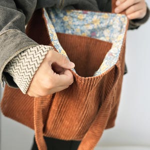 May include: A corduroy tote bag in a warm brown hue. The bag features a floral patterned lining in shades of blue, yellow, and white. The bag has a rectangular shape with a large open compartment and long straps for carrying over the shoulder.