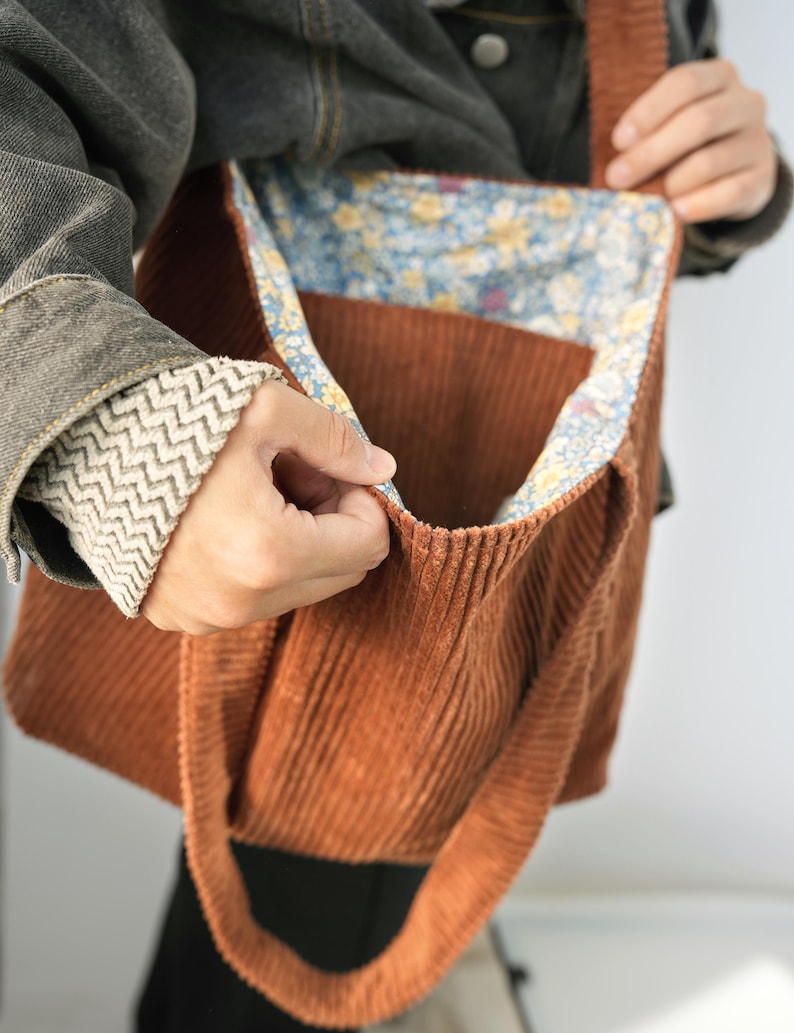 May include: A corduroy tote bag in a warm brown hue. The bag features a floral patterned lining in shades of blue, yellow, and white. The bag has a rectangular shape with a large open compartment and long straps for carrying over the shoulder.