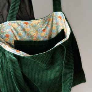 Peut inclure: Un sac fourre-tout en velours côtelé vert foncé avec des sangles assorties. L'intérieur est doublé d'un motif floral avec des fleurs bleues, jaunes et oranges. Le sac a une grande poche à l'avant. Le sac est conçu pour transporter des objets.
