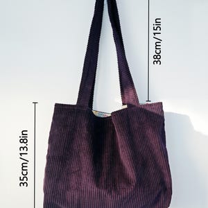 Op de afbeelding: Een pruimkleurige corduroy tote bag met een rechthoekige vorm. De tas is 35 cm breed, 35 cm diep en 38 cm hoog. De tas heeft twee lange hengsels en hangt tegen een witte muur.