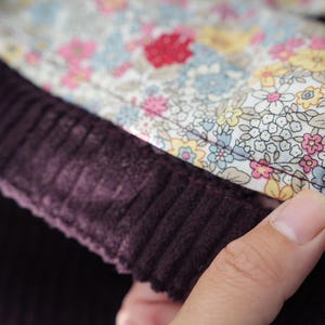 Op de afbeelding: Close-up van een stuk stof. De afbeelding toont een donkerpaars corduroy materiaal naast een bloemmotiefstof. De bloemenstof heeft een witte achtergrond met roze, gele en blauwe bloemen. De stoffen zijn aan elkaar genaaid.