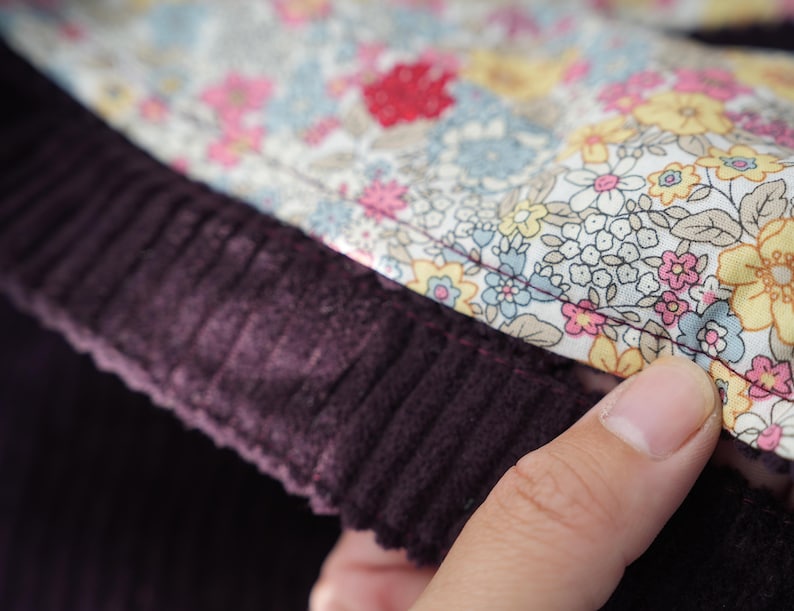 Op de afbeelding: Close-up van een stuk stof. De afbeelding toont een donkerpaars corduroy materiaal naast een bloemmotiefstof. De bloemenstof heeft een witte achtergrond met roze, gele en blauwe bloemen. De stoffen zijn aan elkaar genaaid.