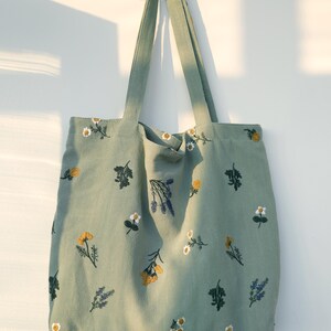 May include: A sage green tote bag with embroidered floral designs. The bag features various flowers in yellow, white, and purple. The tote has long straps for carrying and is hanging against a white wall.
