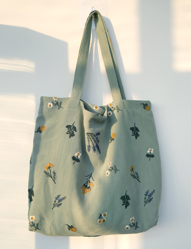 May include: A sage green tote bag with embroidered floral designs. The bag features various flowers in yellow, white, and purple. The tote has long straps for carrying and is hanging against a white wall.