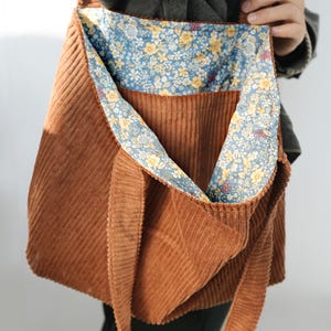 May include: A brown corduroy tote bag with a floral patterned lining. The bag has a long strap for over-the-shoulder or crossbody wear. The interior lining features a blue background with yellow and white flowers.