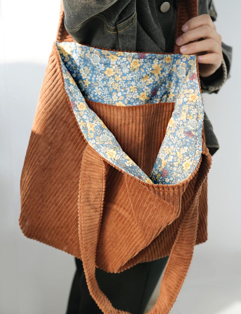 May include: A brown corduroy tote bag with a floral patterned lining. The bag has a long strap for over-the-shoulder or crossbody wear. The interior lining features a blue background with yellow and white flowers.