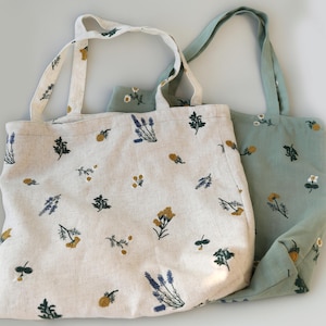 May include: Two tote bags with floral embroidery. One bag is off-white with blue, yellow, and green floral designs. The other is light green with similar embroidery. Both bags have matching handles.