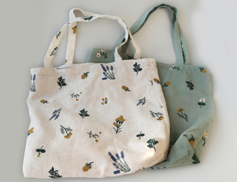 May include: Two tote bags with floral embroidery. One bag is off-white with blue, yellow, and green floral designs. The other is light green with similar embroidery. Both bags have matching handles.