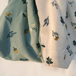 May include: Two fabric pieces, one in a soft green and the other in a natural beige, are adorned with embroidered floral patterns. The designs feature yellow, white, and blue flowers, along with green foliage. The fabric appears to be linen or a similar natural material.