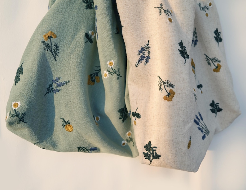 May include: Two fabric pieces, one in a soft green and the other in a natural beige, are adorned with embroidered floral patterns. The designs feature yellow, white, and blue flowers, along with green foliage. The fabric appears to be linen or a similar natural material.
