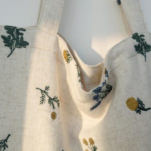 May include: Cream-colored tote bag with embroidered floral designs in shades of green, yellow, and blue. The bag features two sturdy straps and a spacious interior, perfect for carrying everyday essentials. The floral embroidery adds a touch of nature-inspired charm to the bag.