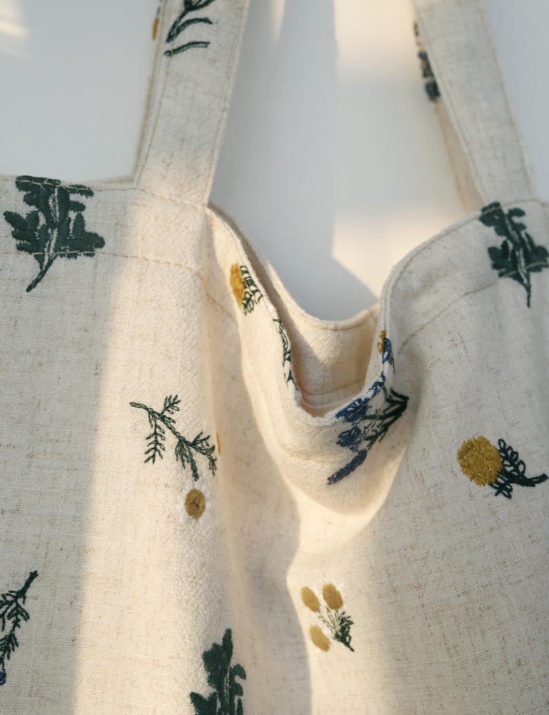 May include: Cream-colored tote bag with embroidered floral designs in shades of green, yellow, and blue. The bag features two sturdy straps and a spacious interior, perfect for carrying everyday essentials. The floral embroidery adds a touch of nature-inspired charm to the bag.
