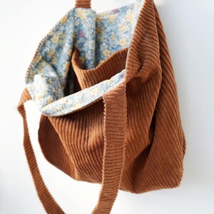 May include: A brown corduroy tote bag with long straps. The interior lining features a blue floral pattern with yellow flowers. The bag is open, revealing the patterned lining and a small pocket.