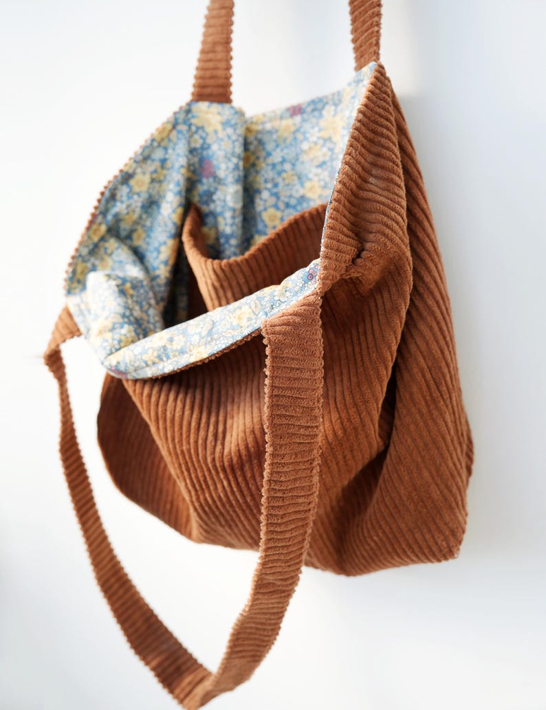 May include: A brown corduroy tote bag with long straps. The interior lining features a blue floral pattern with yellow flowers. The bag is open, revealing the patterned lining and a small pocket.