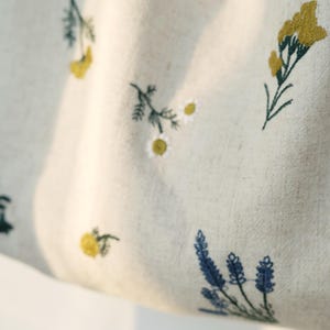 May include: Close-up of a light beige fabric with embroidered floral patterns. The design features yellow, blue, and green flowers and foliage. The fabric has a natural, textured appearance, suggesting a linen or cotton material.