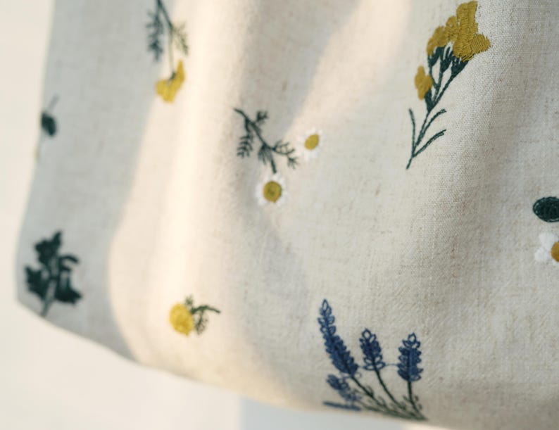 May include: Close-up of a light beige fabric with embroidered floral patterns. The design features yellow, blue, and green flowers and foliage. The fabric has a natural, textured appearance, suggesting a linen or cotton material.