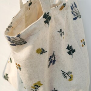 May include: Cream-colored tote bag with embroidered floral designs in yellow, blue, and green. The bag has two handles and a spacious interior. The fabric appears to be linen or a similar natural material, perfect for carrying groceries or personal items.