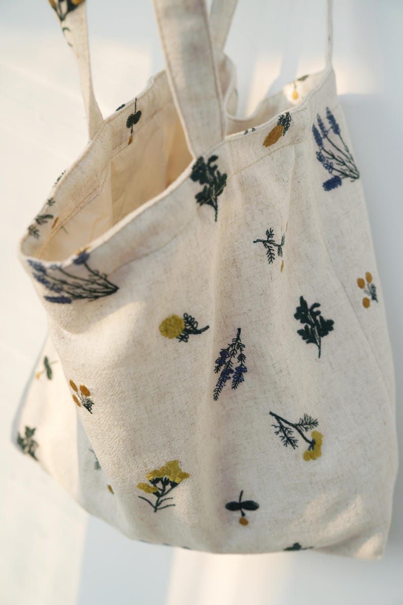 May include: Cream-colored tote bag with embroidered floral designs in yellow, blue, and green. The bag has two handles and a spacious interior. The fabric appears to be linen or a similar natural material, perfect for carrying groceries or personal items.