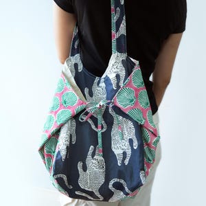 May include: A fabric tote bag featuring a dark blue panel with a white tiger print and side panels in pink with a green seashell pattern. The bag includes a matching fabric tie closure at the top center and two shoulder straps for carrying.