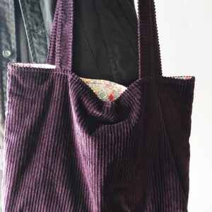 Op de afbeelding: Een dieppaarse corduroy tote bag met lange hengsels. De binnenvoering van de tas heeft een bloemenpatroon met rode en gele accenten. De tas is gemaakt van een zachte, geribbelde stof.