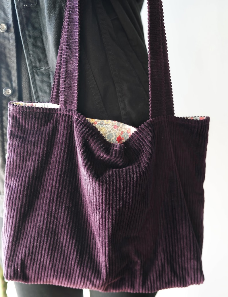 Op de afbeelding: Een dieppaarse corduroy tote bag met lange hengsels. De binnenvoering van de tas heeft een bloemenpatroon met rode en gele accenten. De tas is gemaakt van een zachte, geribbelde stof.