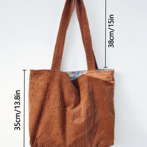 May include: A brown corduroy tote bag with a floral patterned lining. The bag measures 13.8 inches wide, 13.8 inches high, and has 15-inch-long handles. The bag is made of a soft, ribbed fabric and is designed for carrying items.
