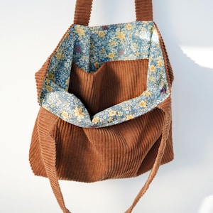 May include: A brown corduroy tote bag with a floral patterned lining. The bag has a rectangular shape with a large open compartment and two long straps. The interior lining features a blue background with yellow and white flowers.
