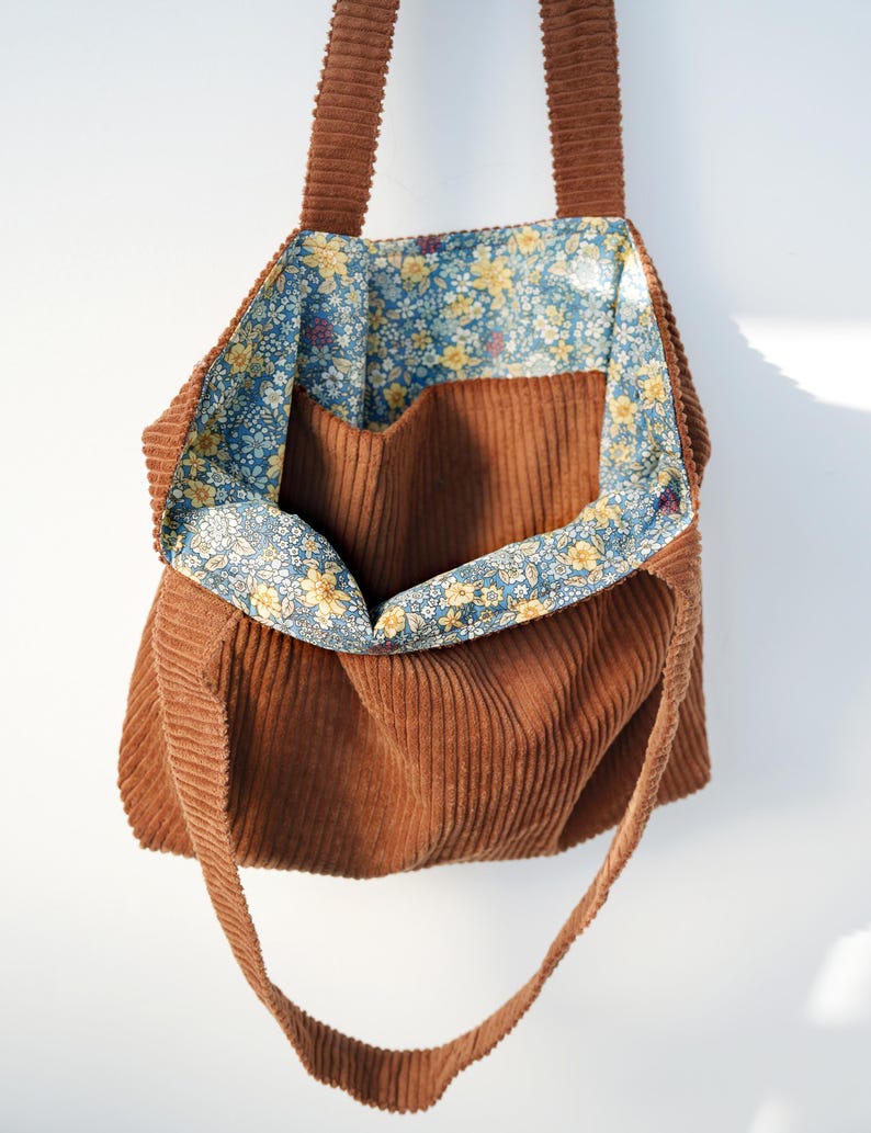 May include: A brown corduroy tote bag with a floral patterned lining. The bag has a rectangular shape with a large open compartment and two long straps. The interior lining features a blue background with yellow and white flowers.