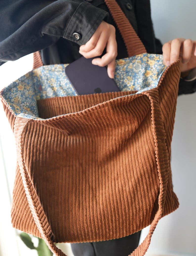 May include: A brown corduroy tote bag with a floral patterned lining. The bag has a rectangular shape with a large main compartment and a smaller pocket. The handles and exterior are a solid brown color. A person is putting a phone into the pocket.